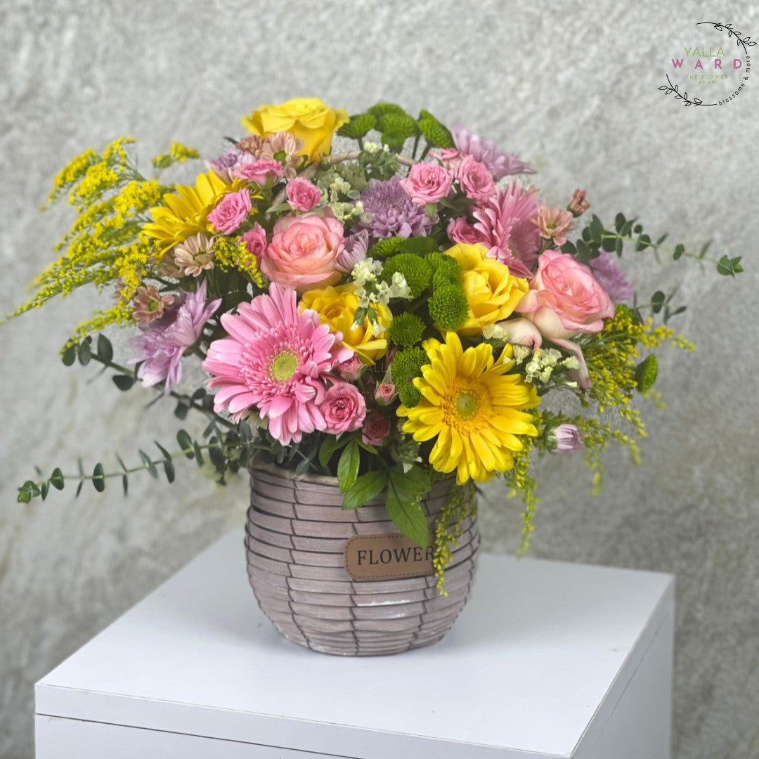  colorful flowers in a woven basket on a white surface with a gray textured background.