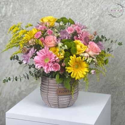  colorful flowers in a woven basket on a white surface with a gray textured background.
