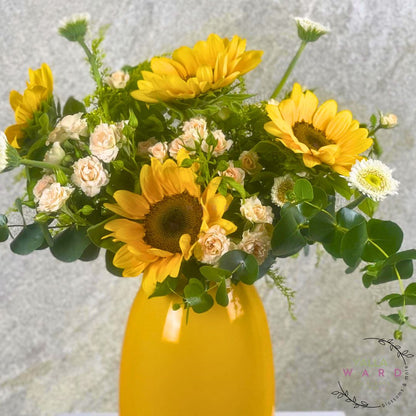 Bouquet of sunflowers and small white flowers in a yellow vase against a light gray background.