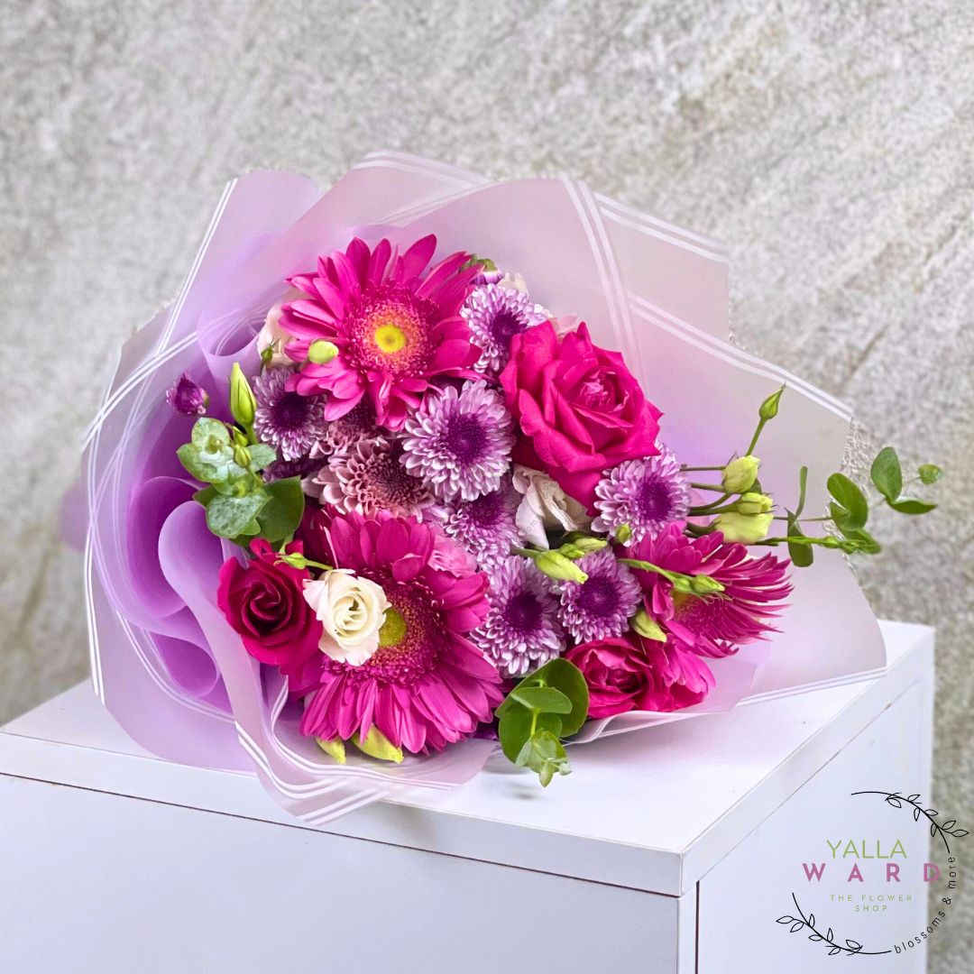 Bouquet of pink and purple flowers wrapped in clear plastic on a white surface with a light gray textured background.
