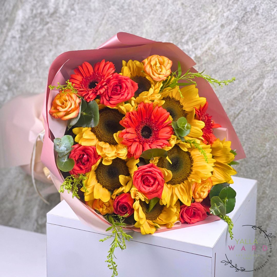 Bouquet of sunflowers, yellow, and orange flowers in a pink wrap on a white surface with a light gray background.