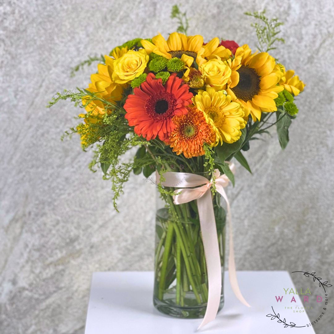 Bouquet of flowers with sunflowers, roses, and daisies in a clear vase on a textured surface.