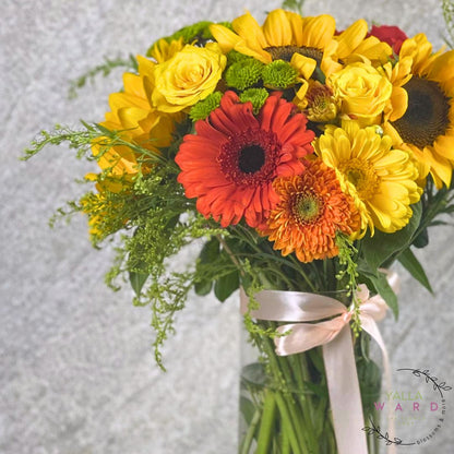 Bouquet of flowers with yellow sunflowers, red and orange gerberas, and greenery on a light gray background.