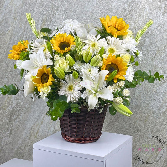 yellow and white flowers in a woven basket on a white pedestal against a gray stone wall.