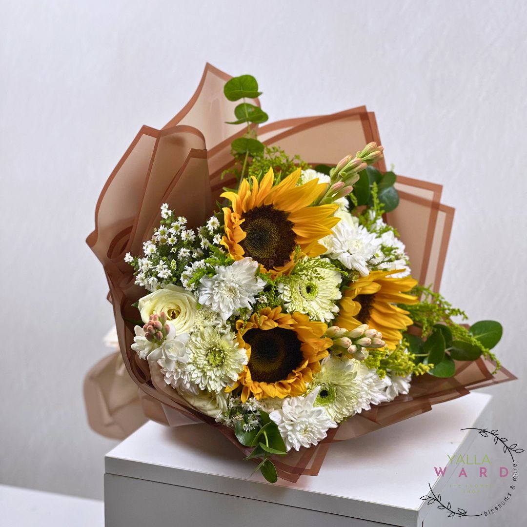 Bouquet of sunflowers and white flowers wrapped in brown paper on a white surface with a light gray background.