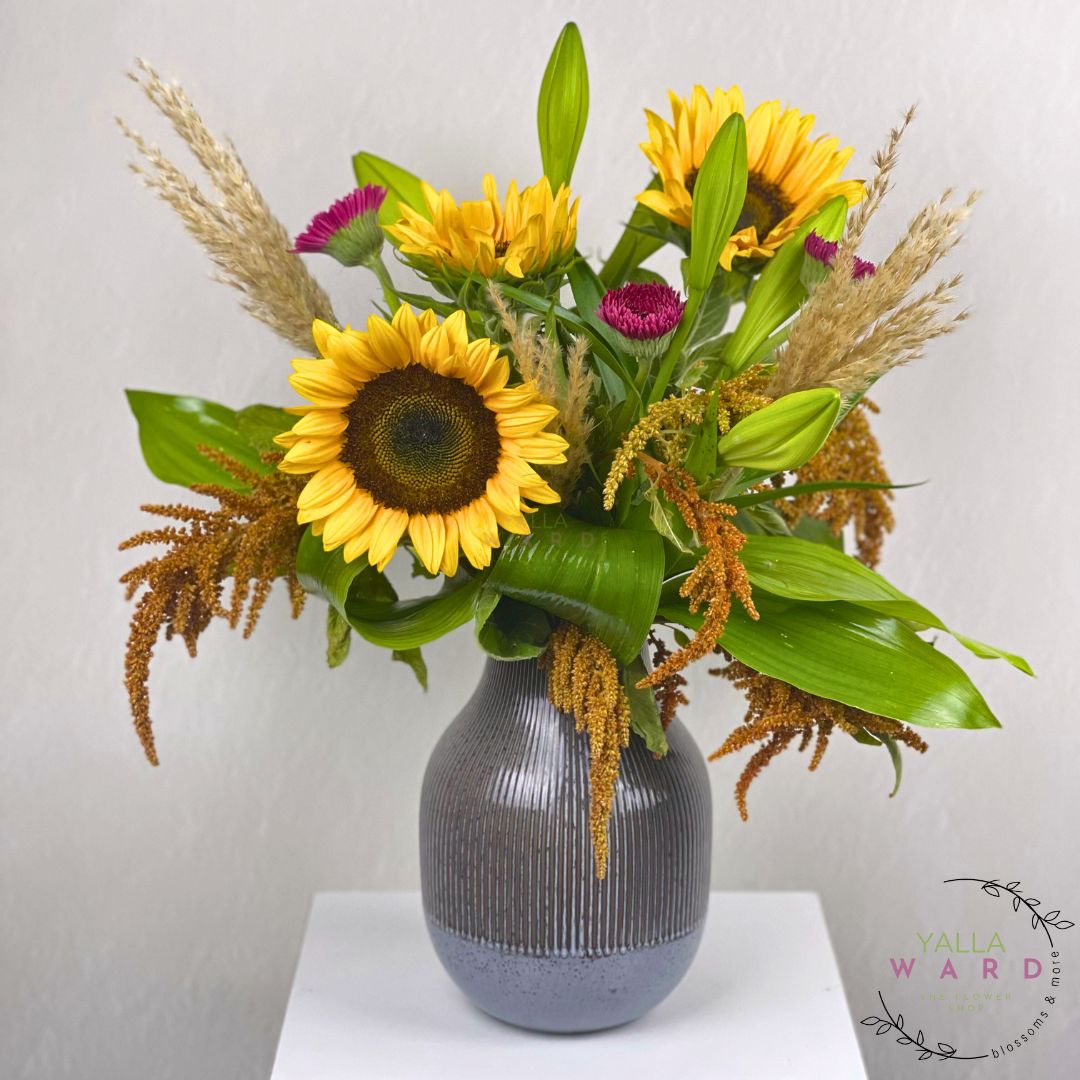 Floral arrangement with sunflowers and greenery in a ribbed gray vase on a white surface.