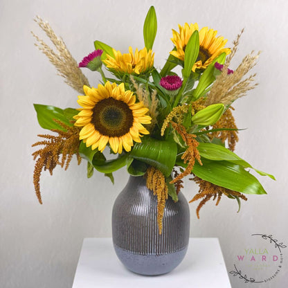 Floral arrangement with sunflowers and greenery in a ribbed gray vase on a white surface.