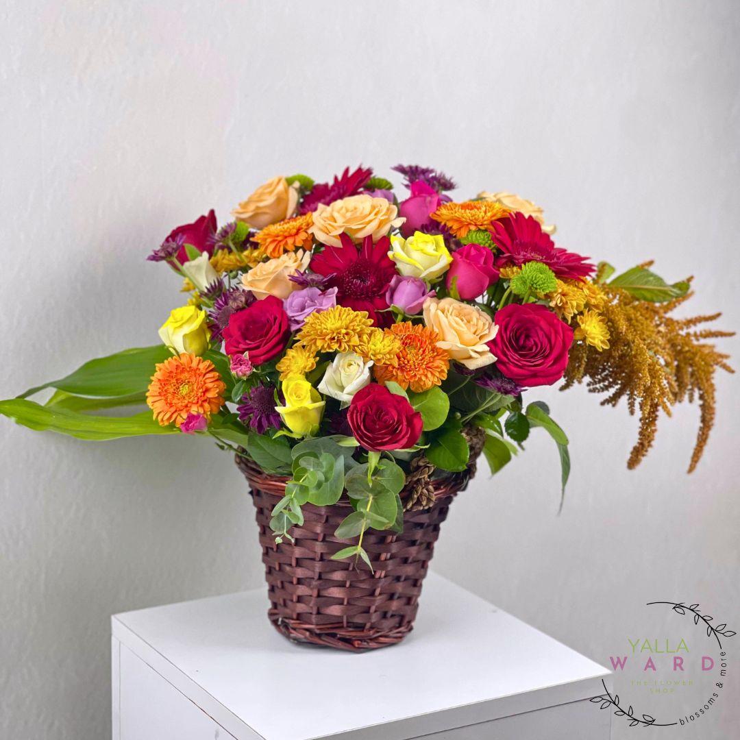 Colorful flower arrangement in a wicker basket on a white surface with a light gray background.