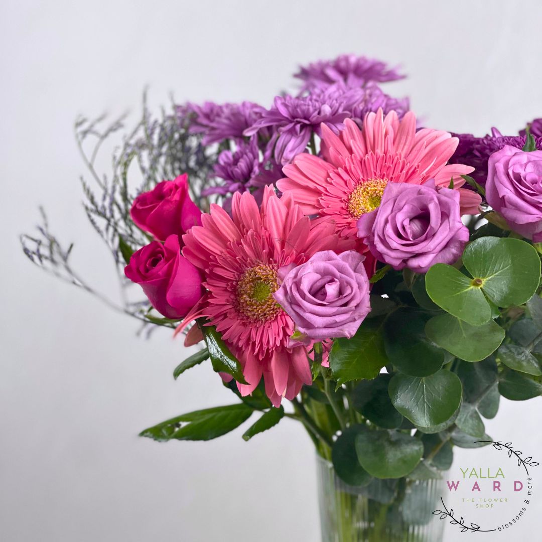 pink and purple flowers in a clear vase on a light gray background with 'Yalla Ward' branding.