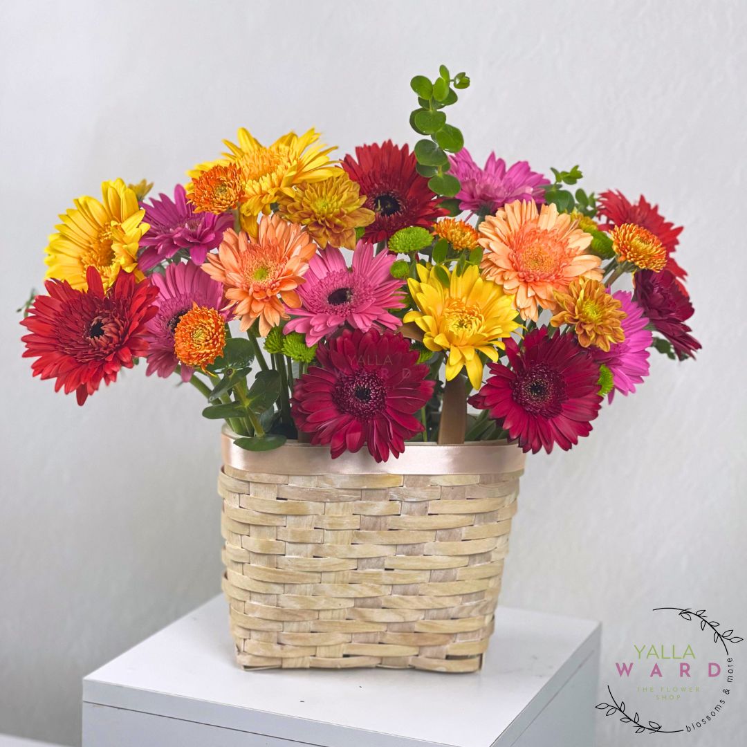 Colorful bouquet of flowers in a woven basket on a white surface with a light gray background.