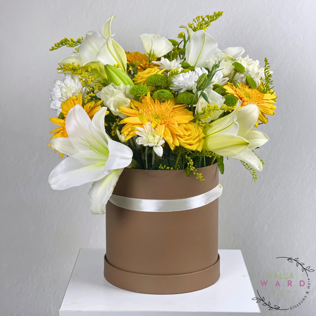 Floral arrangement in a brown box with yellow and white flowers on a white surface.