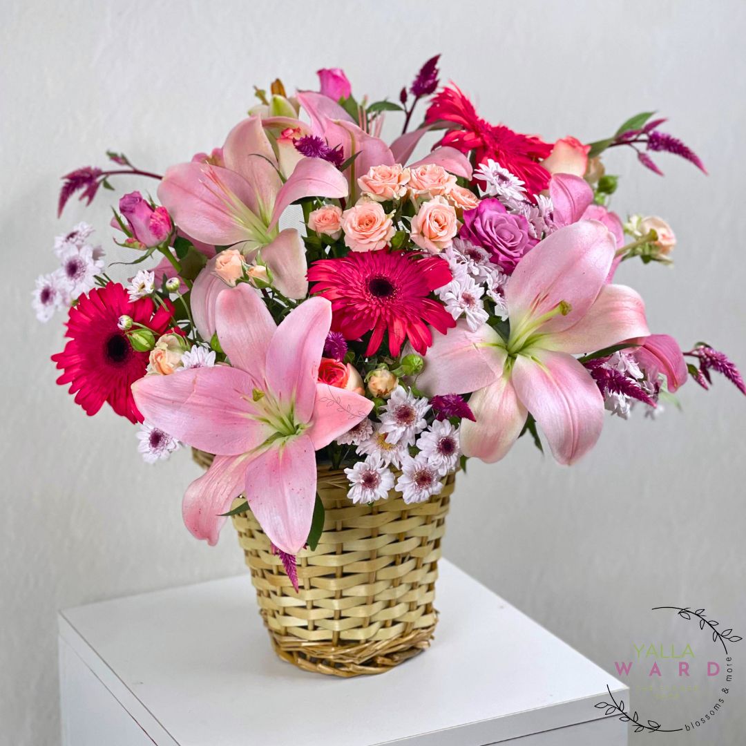 pink lilies and other flowers in a woven basket on a white surface with a light gray background.