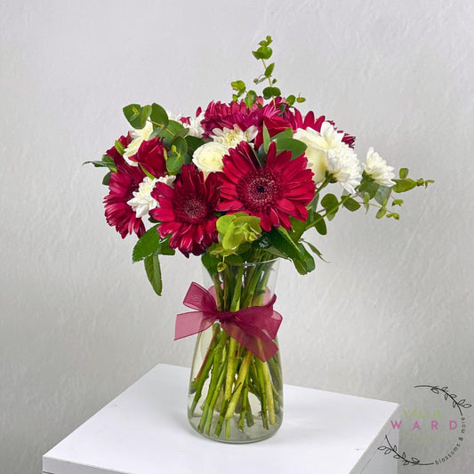 red, white, and green flowers in a clear vase with a pink ribbon on a white surface.
