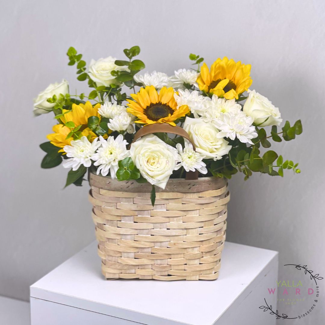 Floral arrangement with sunflowers and white flowers in a woven basket on a white surface.