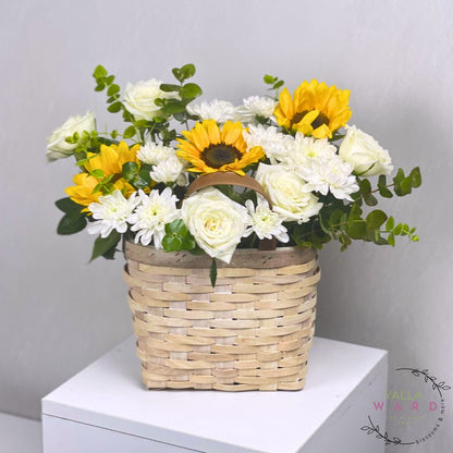 Floral arrangement with sunflowers and white flowers in a woven basket on a white surface.