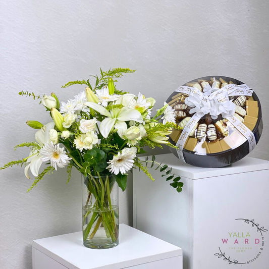 Bouquet of white flowers in a vase next to a chocolate gift on a white surface with a light gray background.
