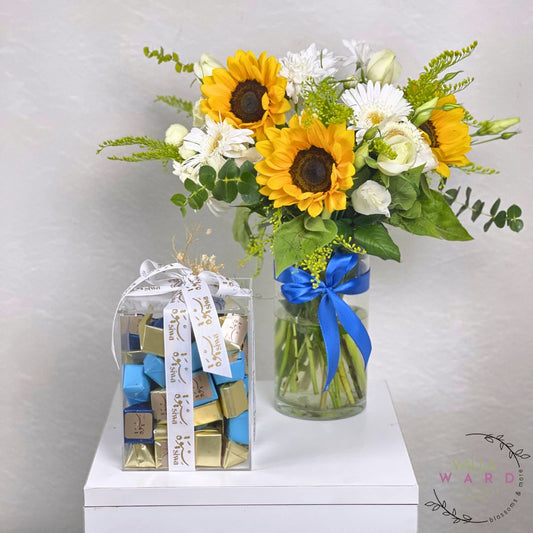 Bouquet of sunflowers and white flowers in a glass vase with a blue ribbon, next to a transparent chocolate box 