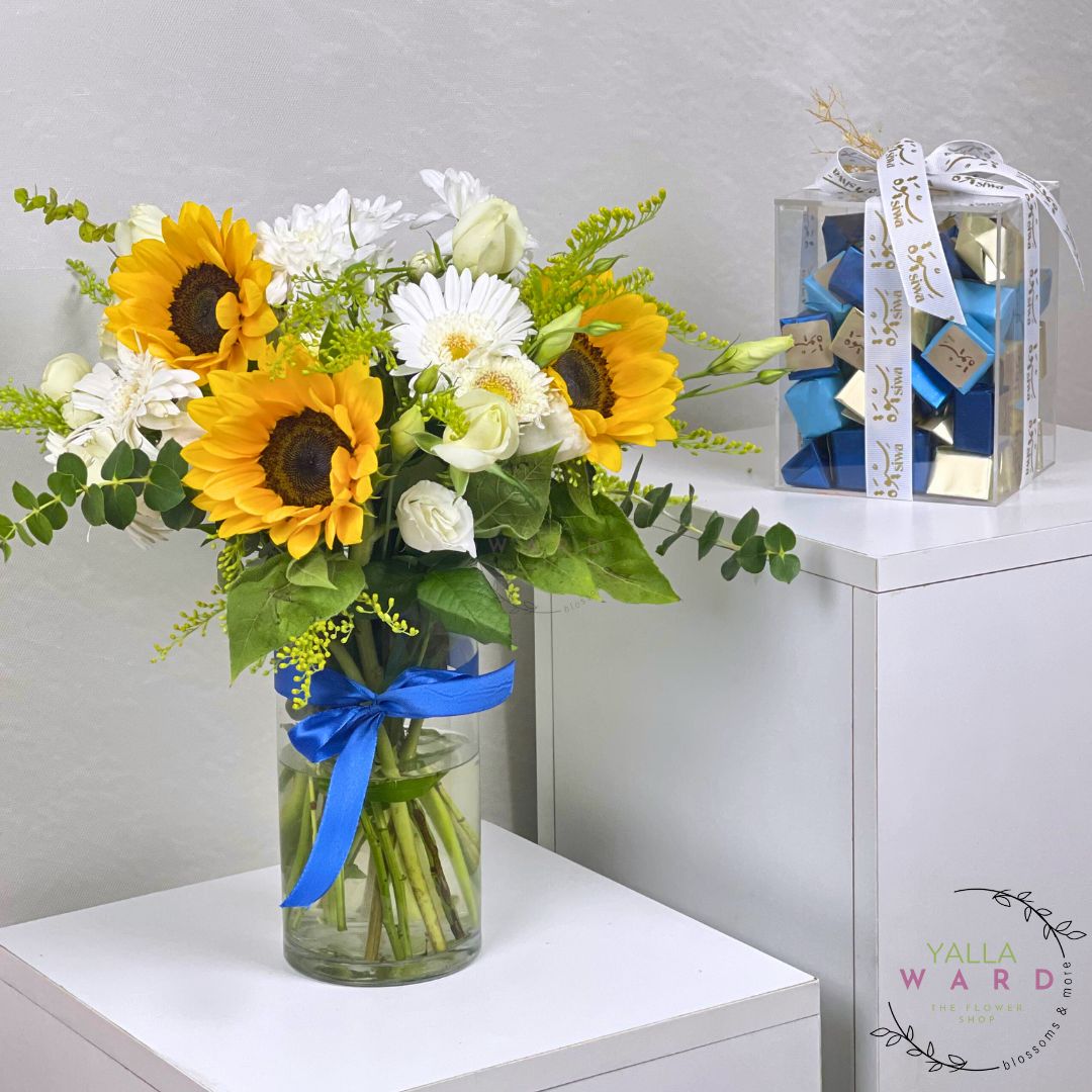 Bouquet of sunflowers and white flowers in a clear vase with a blue ribbon, placed on a white surface. with chocolate box 
