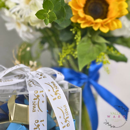 Bouquet of flowers with a blue ribbon and clear box, featuring a brand logo.