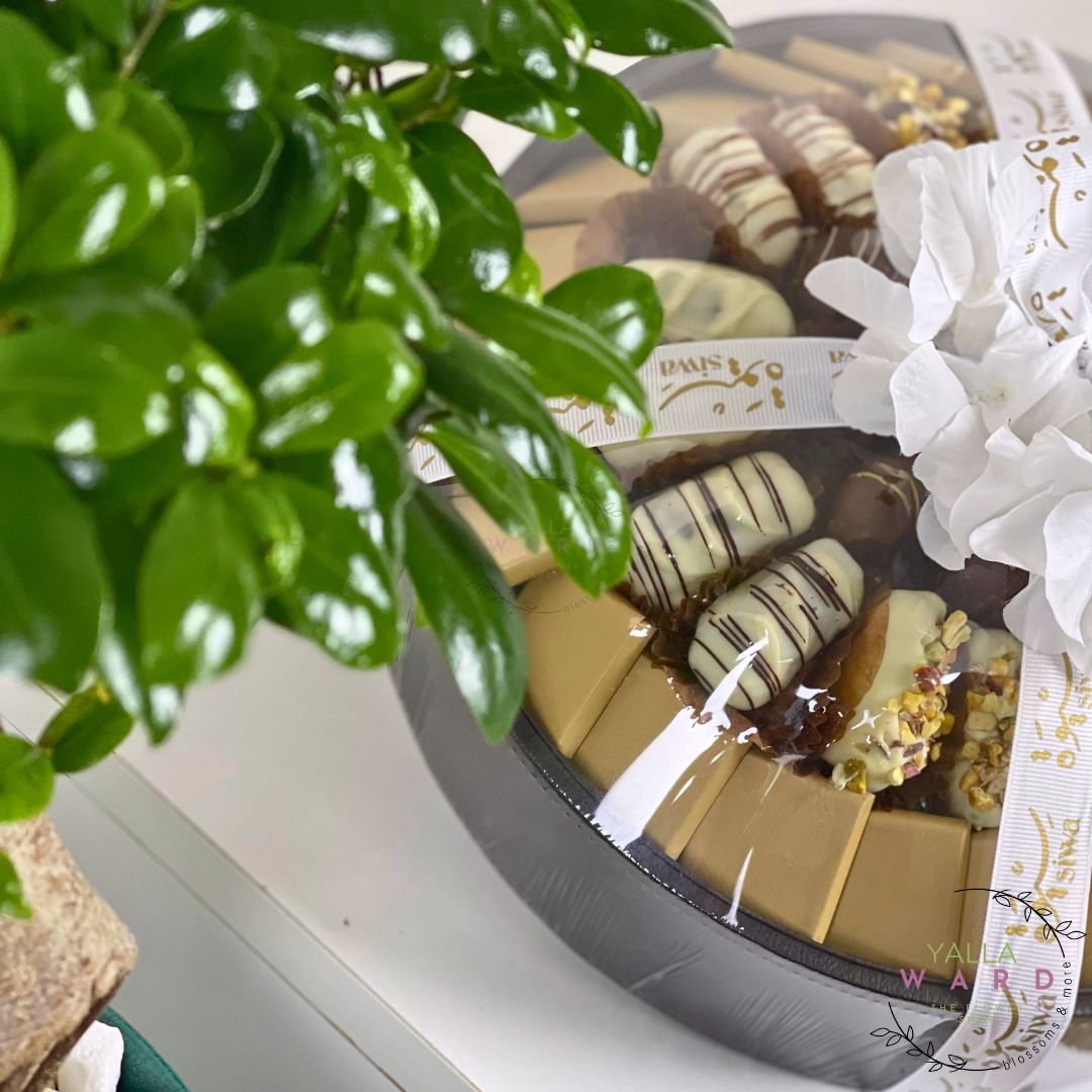 Box of chocolates with a decorative ribbon on a white surface, surrounded by green leaves.