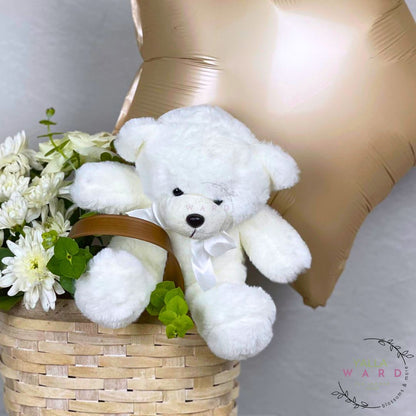 White teddy bear in a basket with flowers and a gold balloon on a light gray background