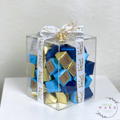 Clear gift box with blue and gold chocolate bars inside, tied with a white ribbon.