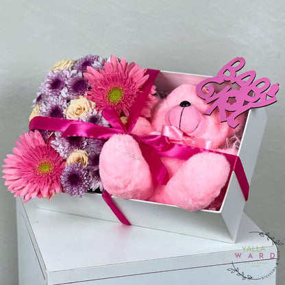 Pink teddy bear and flowers in a box with 'Love' sign on a white surface