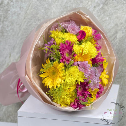 Bouquet of yellow and pink flowers in a pink wrap on a white surface with a light gray background.