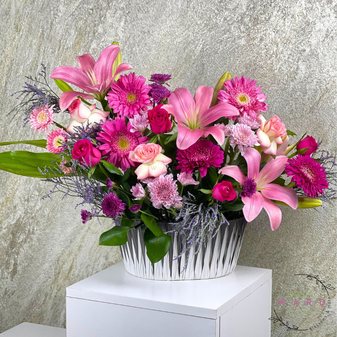  pink and purple flowers in a silver basket on a white surface with a textured gray background.